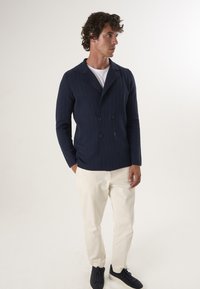 Marineblauwe dubbelbreasted jas met een gestructureerd patroon, voorzien van een sjaalkraag en twee knopen, gecombineerd met lichtbeige broek en donkere schoenen.