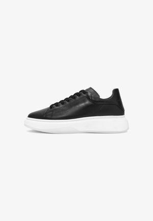 Zapatilla de cuero negro con puntera redondeada, suela texturizada blanca y seis ojales para los cordones. Diseño minimalista, superficie lisa.