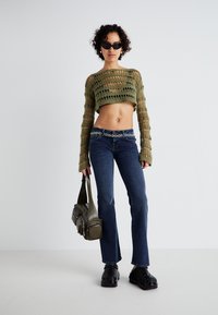 Grønn strikket crop top med lange ermer, blå jeans og sorte klumper. Tilbehøret inkluderer et mønstret belte og en grønn skulderveske.
