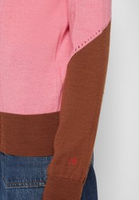 Manche de pull en maille à blocs de couleurs rose et marron avec les initiales "VB" brodées en rouge, portée avec un jean en denim bleu.