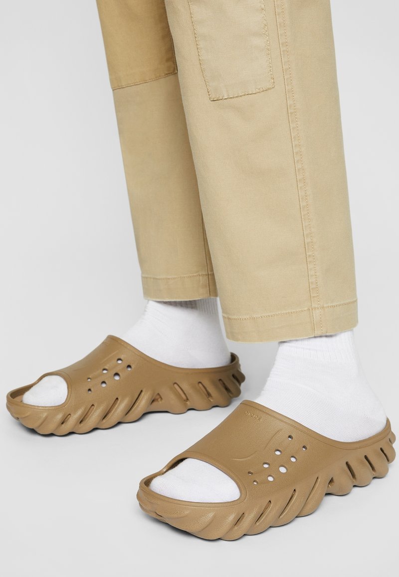 Sandales diapositive moulées marron avec un orteil ouvert, des côtés perforés et un design de semelle ondulée. Associées à des chaussettes blanches et un pantalon khaki.