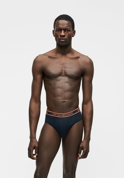 Μυώδης μαύρος άνδρας στέκεται προς τα εμπρός φορώντας σκούρα μπλε ανδρικά εσώρουχα Emporio Armani με πορτοκαλί ζώνη μέσης σε απλό φωτεινό φόντο.