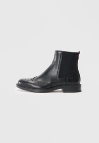 Stivale Chelsea in pelle nera con dettagli a brogue, pannelli laterali elastici e una linguetta sul retro. Tacco basso e design elegante.