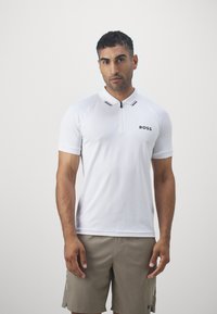 White polo shirt with a zippered collar, black logo, and subtle striped accents on the collar, paired with beige shorts. Smooth texture.