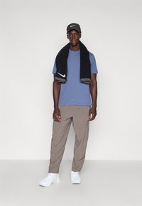 Compleu sport Nike, constând dintr-un tricou albastru, o șapcă gri, un prosop negru și pantaloni scurți bej. Tenisii albi completează look-ul.