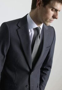 Chaqueta de traje azul marino oscuro con solapas muesca y dos botones. Camisa de vestir blanca debajo, complementada por una corbata gris texturizada.