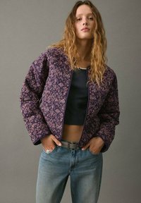 Giacca bomber trapuntata viola con motivo floreale, bordi blu navy e vestibilità rilassata. Indossata sopra un top corto scuro e abbinata a jeans chiari.