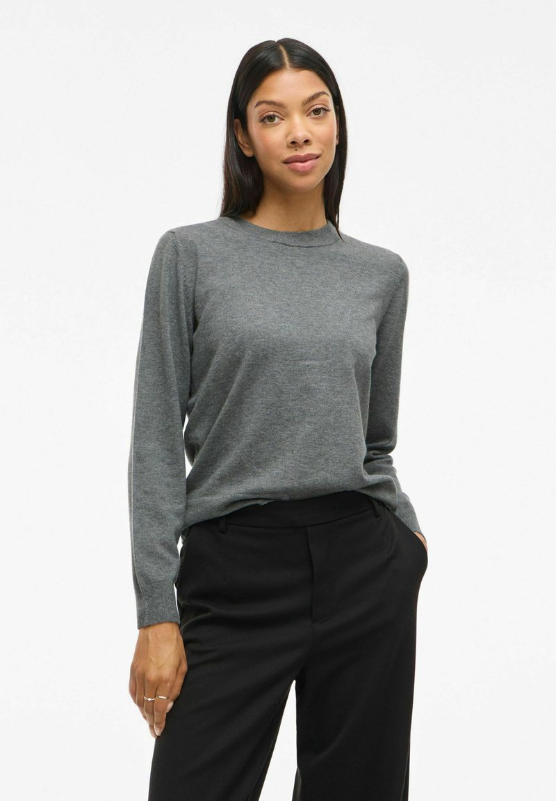 Pull gris à manches longues en tissu doux, avec un col rond et des poignets ajustés, associé à un pantalon noir ajusté.