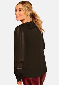 Blonde vrouw met een paardenstaart draagt een doorzichtige zwarte blouse met een gerimpelde kraag en gedetailleerde mouwen, gecombineerd met donkerrode leren broek.