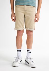 Pantalones cortos beige con dobladillos, hechos de material vaquero. Combinados con zapatillas blancas y calcetines con dos rayas negras.