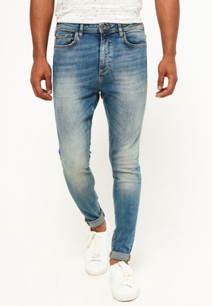 Man draagt lichtblauwe slim-fit jeans met omgeslagen zomen en witte sneakers, staand tegen een witte achtergrond.