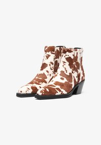 Καφέ και λευκά μποτάκια καουμπόισσας, φτιαγμένα από υφή γούνας, με μυτερή μύτη και χοντρό μαύρο τακούνι. Κομψός σχεδιασμός με ελαστικές πλαϊνές πάνελ.