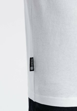 Biała bawełniana koszulka z krótkim rękawem, z czarną metką marki umieszczoną na bocznym szwie. Gładka tekstura i prosty design.