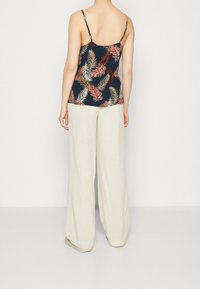 Nainen, jolla on yllään tummansinisestä kankaasta valmistettu toppi, jossa on monivärisiä lehtikuvioita, ja hameet, jotka ovat löysät beige-väriset housut, seisoo yksinkertaista valkoista taustaa vasten.