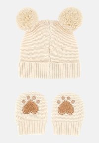 Cappello per neonati lavorato a maglia di colore crema con due grandi pompon e muffole coordinate che presentano disegni di cuori e impronte di zampe marroni.