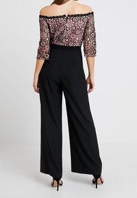 Un pantalon large noir associé à un haut en dentelle florale ajusté, dégageant les épaules. Le haut présente des manches trois-quarts et un ourlet en pointes.