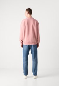 Sudadera oversized rosa claro con hombros caídos, combinada con jeans de mezclilla azul. Se llevan zapatillas blancas, con una textura suave.