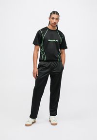 Čierny športový outfit pozostávajúci z trička s krátkym rukávom so zelenými akcentmi a logom, v kombinácii s čiernymi jogger nohavicami a bielymi teniskami.