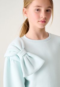 Lichtblauwe sweatshirt met een grote strikaccent aan één schouder. Zachte stof, ronde halslijn en geplooide mouwdetails.