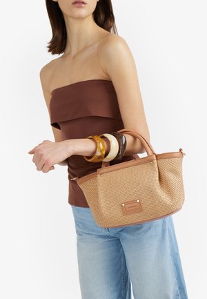 Donna che indossa un top marrone senza spalline e jeans blu, con in mano una borsa beige intrecciata e tre braccialetti spessi in resina al polso.