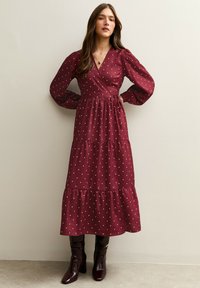 Bordeauxrotes Wickelkleid mit weißen Polka-Dots, langen Puffärmeln, gestuftem Rock und Taillenband, kombiniert mit kniehohen braunen Stiefeln.