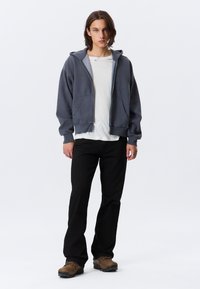 Grå hettejakke med frontlommer, båret over en hvit skjorte. Svarte bukser og brune sko fullfører antrekket. Enkelt, uformelt design.