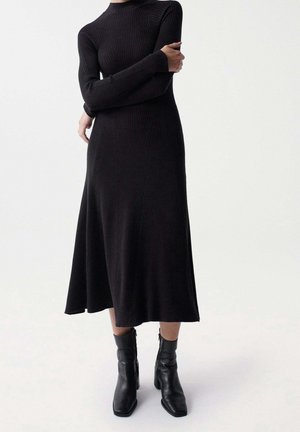 Mujer vestida con un vestido largo de punto acanalado negro con mangas largas y botines negros de tacón, de pie con los brazos cruzados.