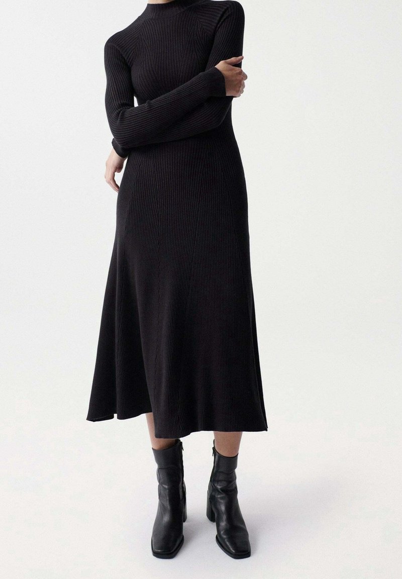 Femme portant une longue robe noire en maille côtelée à manches longues et des bottines à talons noires, debout les bras croisés.
