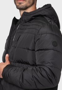 Doudoune noire avec capuche, dotée d'une poche zippée sur la poitrine, d'une texture matelassée et d'un écusson rond sur l'épaule.