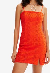 Robe rouge sans manches avec des motifs floraux brodés, silhouette ajustée, fente cuisse et décolleté carré. Accentuée par un collier cœur en argent.