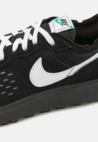 Svart sneakers med ovandel i mocka och mesh, vit Nike-logotyp, vita snören och en strukturerad svart sula för bra grepp.