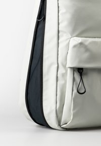 Zaino grigio chiaro con cerniera nera, dotato di un pannello laterale testurizzato e una tasca frontale con cordino. Realizzato in materiale resistente.