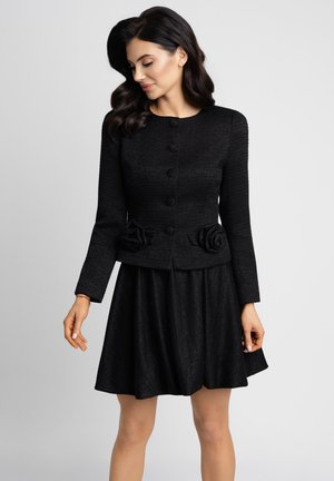 Kobieta ubrana w czarną, fakturowaną marynarkę z długim rękawem z kwiatowymi zdobieniami z tkaniny oraz dopasowaną rozkloszowaną spódnicę, stojąca na jednolitym tle.