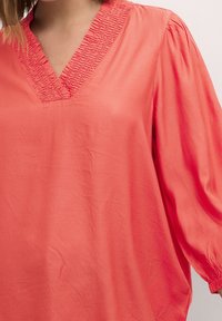 Blusa corallo con scollatura a V e dettagli arricciati lungo il collo. Presenta maniche lunghe e voluminose e una superficie liscia e opaca.