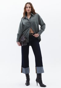 Camicia grigia a maniche lunghe con bottoni e tasca frontale. Pantaloni a campana in denim scuro con risvolti in denim a contrasto. Stivali neri con tacco alto. Borsa marrone.