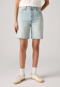 Ανοικτού μπλε τζιν σορτσάκια με ψηλή μέση, πέντε τσέπες και σαγρέ άκρη. Φοριούνται με λευκά παπούτσια και ριγέ κάλτσες.