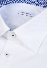 Col de chemise blanche texturée avec doublure intérieure à motif bleu et deux boutons blancs avec coutures bleues, étiquette de marque "seidensticker" visible.