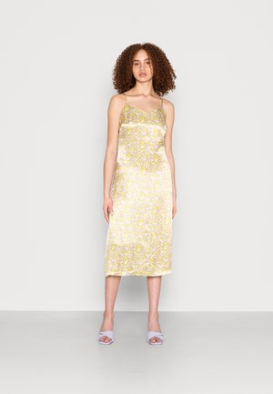 Vestido de satin floral com alças finas, apresentando um padrão amarelo e branco. Corte a meio comprimento com uma textura suave e brilhante. Combinado com sapatos lilases claros.