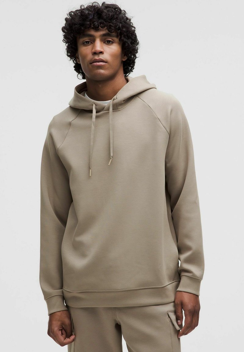 Mlad moški s kodrastimi lasmi, oblečen v svetlo rjav hoodie, stoji pred enostavno ozadje in gleda neposredno v kamero.