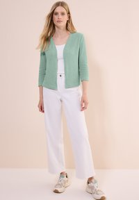 Cardigan texturé vert menthe avec des manches trois quarts, associé à un débardeur blanc et un pantalon large blanc ; porté avec des baskets beiges et vertes.
