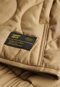 Gequilte beige stof met een textuurafwerking. Voorzien van een zwart label met gele tekst met de details "Superdry Surplus" en onderhoudsinstructies.