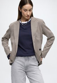 Hnedo-béžové kárované sako s výrezovým golierom cez tmavomodrý sveter, doplnené svetlosivými džínsami; vyrobené z textúrovanej látky.