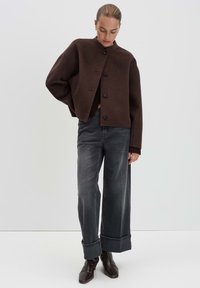 Vrouw in een donkerbruine knopenjack, met brede, opgerolde grijze jeans en donkerbruine vierkante laarzen, staand tegen een witte achtergrond.