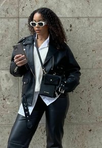 Schwarze Lederjacke mit silbernen Beschlägen, über einem weißen Hemd und schwarzen Hosen getragen. Als Accessoire dient eine kleine schwarze Tasche mit einem Riemen.