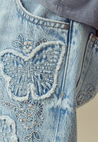 Ljusblå denimjacka med detaljerad broderi av en fjäril och blommor, med fransade kanter och en bleknad textur.