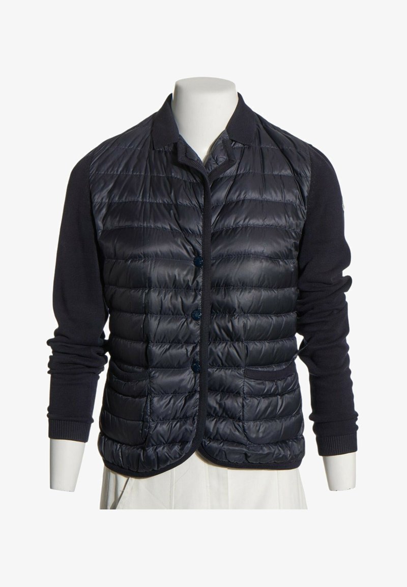 Gilet matelassé rembourré bleu marine avec fermeture par boutons, porté sur un haut en maille noir à manches longues, sur un buste de mannequin blanc.
