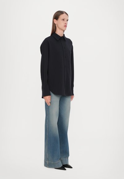 Camicia nera con colletto, abbinata a jeans sbiaditi a vita alta e gamba larga, e scarpe nere a punta. Texture del tessuto morbido.