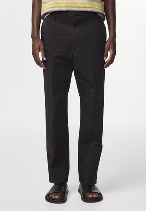 Schwarze Baumwollhose mit geradem Bein, ausgestattet mit einem Knopfverschluss und Taschen. Getragen mit schwarzen Rutschsandalen.