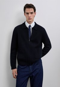 Marineblauwe wollen zip-up cardigan met een kraag, twee voorzakken en geribbelde manchetten. Gedragen over een wit shirt en een blauwe stropdas.