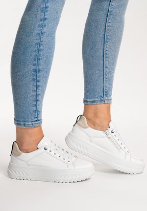 White platform sneakers with beige heel accents, worn with light blue skinny jeans, shown on feet against a plain background.